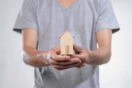 A person in a gray t-shirt presents a miniature wooden house model with both hands, symbolizing home ownership and new beginnings, capturing a moment of creativity and warmth.の写真素材