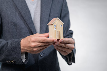A person in a business suit holds a small wooden house model against a neutral grey background, symbolizing real estate investment and the dream of home ownership.の写真素材