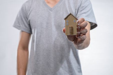 A male hand holds a small wooden house model, symbolizing home and investment. The gray background enhances the model's charm, representing dreams of ownership.の写真素材