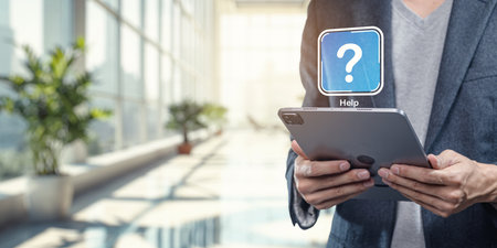 A professional individual stands in a bright office holding a tablet with a help icon displayed, symbolizing support and digital assistance in modern workplaces.の写真素材