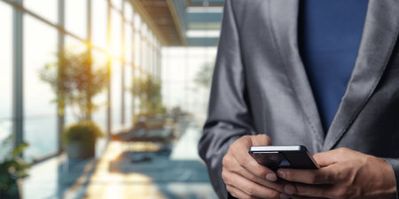 A businessman in a sleek suit engages with his smartphone in a contemporary office. Sunlight streams through large windows, creating a productive atmosphere.の写真素材