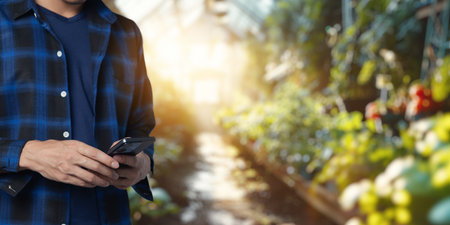 A person in a flannel shirt checks their smartphone in a modern greenhouse filled with vibrant plants, showcasing the integration of technology in business agriculture.の写真素材