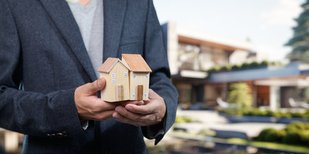 A professional dressed in a suit holds wooden model houses, symbolizing real estate sales and property investment in a modern urban environment.の写真素材
