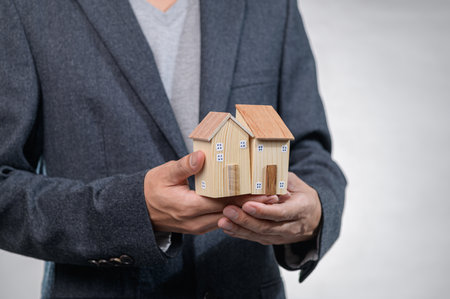 A professional individual in a suit holds two wooden model houses, symbolizing real estate investment and ownership. The setting conveys elegance and ambition.の写真素材
