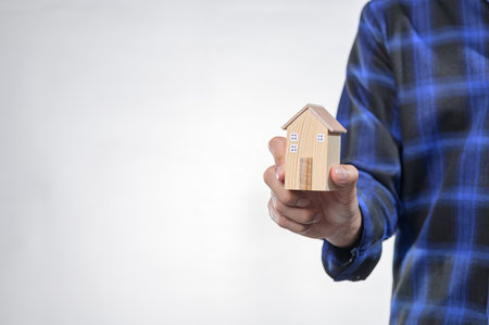 A person holds a small wooden model house symbolizing home ownership and real estate investment. The neutral background emphasizes the model's design.の写真素材