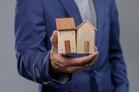 A businessman with a stylish suit holds two miniature wooden houses, symbolizing investment opportunities in the real estate market and housing industry.の写真素材