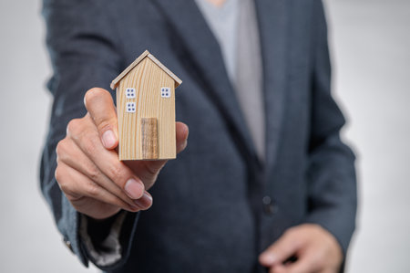 A person in business attire holds a wooden model of a house, symbolizing real estate investment and housing concepts. Ideal for finance or architecture promotions.の写真素材