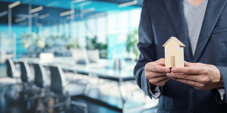 A business professional stands in a modern office, holding a small model house, symbolizing real estate sales and home investment. This image highlights the relationship between business and housing markets.の写真素材