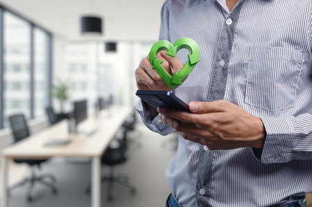 A man stands in a modern office, using a smartphone with a green heart symbol, representing commitment to eco-friendly solutions and sustainable business practices.の写真素材