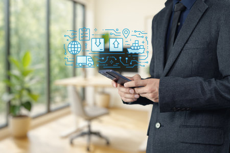 A businessman stands in a modern office using a smartphone. Digital icons representing export and logistics float above his device, symbolizing innovation in business.の写真素材