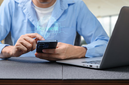 A man engages in a digital payment using a smartphone while seated at a modern desk with a laptop, highlighting the ease of online transactions in today's tech-savvy world.の写真素材