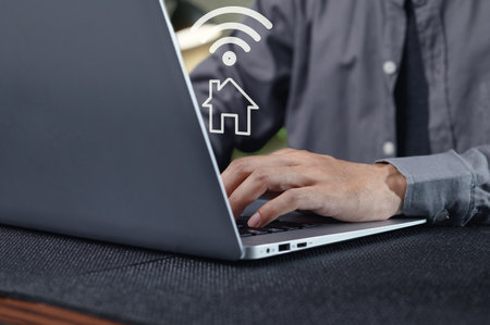 A close-up view of hands typing on a laptop in a home office. The image features a wireless internet connection symbol, emphasizing remote work importance.の写真素材