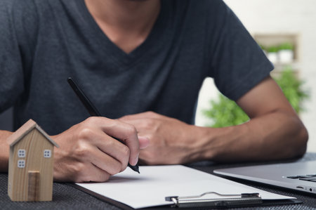 An individual focused on writing notes on a blank paper while a miniature house model sits nearby. This setting reflects themes of property trading and investment decisions in real estate.の写真素材