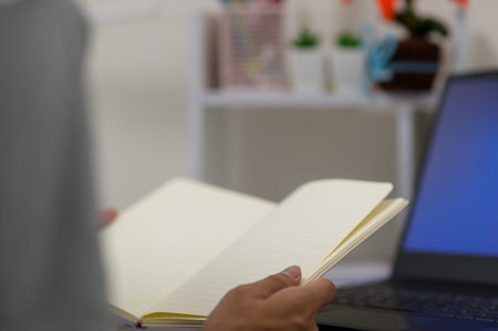 A person is holding an open notebook while sitting in front of a laptop on a desk. The setting...
