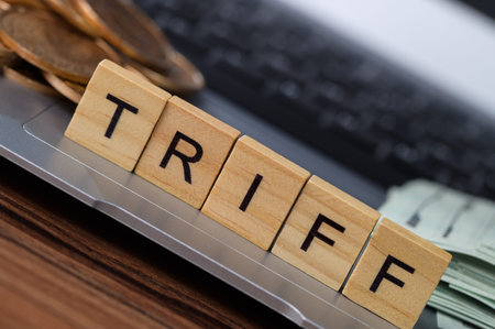 A close-up of wooden scrabble tiles spelling tariff placed on a laptop keyboard, surrounded by coins...