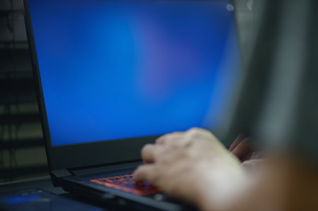 A person is typing on a laptop with an empty blue screen in a dimly lit environment. The image...