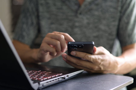 A person engages with a smartphone while working on a laptop in a modern workspace. The image...