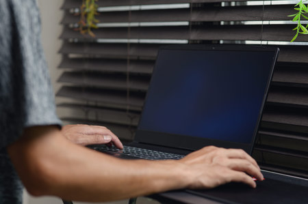 A person engaged with a laptop in a cozy indoor setting, surrounded by window blinds and greenery....