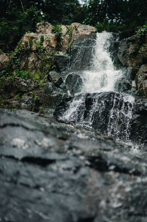 A tranquil waterfall flows gracefully over rocky surfaces, surrounded by lush greenery. This serene scene captures the essence of natural beauty and exploration.の写真素材
