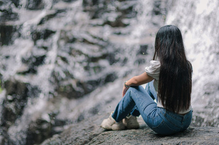 A serene view of a woman sitting near a waterfall, surrounded by nature