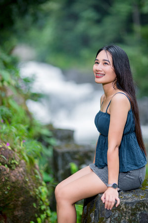 A joyful young woman sits on a stone, surrounded by the lush greenery of a forest, enjoying the tranquil ambiance of a nearby waterfall and stream.の写真素材