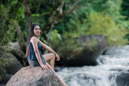 Explore the serene beauty of nature as a girl sits on a rock by a forest stream, surrounded by lush greenery and a gentle waterfall. Perfect for outdoor enthusiasts!の写真素材