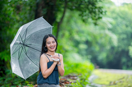 A joyful woman sits in a lush green forest, holding an umbrella. Her smile radiates happiness amidst nature, embodying peace and relaxation in this serene outdoor setting.の写真素材