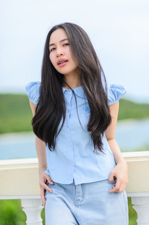 A young woman stands outdoors, radiating confidence and beauty in soft blue attire, with long hair...