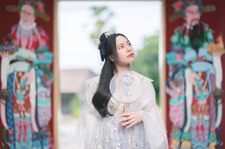 A young woman dressed in traditional Hanfu stands gracefully, holding a decorative fan. The serene...