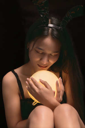 A serene woman in a black bunny costume gently cradles a glowing ball, embodying the playful spirit of Halloween. The soft lighting creates a cozy atmosphere.の写真素材