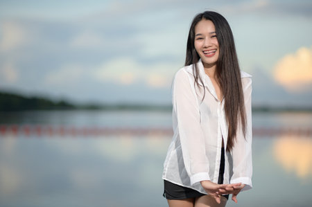 A young woman in a white shirt stands at the beach, smiling joyfully. The calm water and beautiful sky create a serene atmosphere.の写真素材