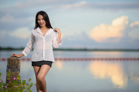 A woman stands gracefully by the water, wearing a sheer white shirt. The beautiful sunset reflects off the calm surface, creating a serene atmosphere.の写真素材