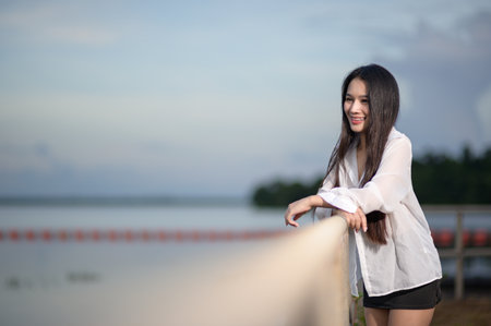 A young woman stands by the water, smiling in casual attire. The serene environment and bright daylight capture a joyful moment in nature, perfect for tourism themes.の写真素材