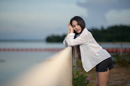 A young woman in a white shirt stands by a tranquil water body, enjoying nature's beauty in a serene tourism setting. An ideal capture of relaxation and charm.の写真素材