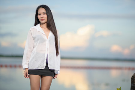 A young woman stands confidently by a tranquil water body during sunset, showcasing her casual style in a white shirt, perfect for nature tourism themes.の写真素材