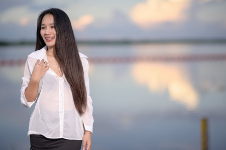 A young woman in a stylish uniform stands by the serene water, smiling under the cloudy sky.の写真素材