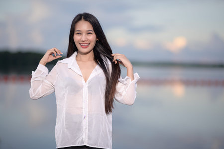 A cheerful young woman poses confidently in front of serene water. Her casual style and joyful...