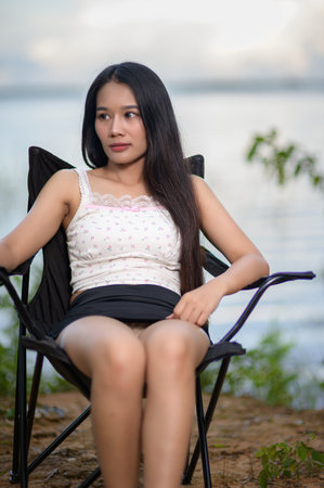 A young woman sits relaxed in a camping chair, enjoying the scenic view of a tranquil reservoir. The natural setting enhances her sense of peace and contentment.の写真素材