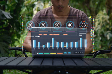 A man works outdoors on his laptop, surrounded by lush greenery. The screen displays dynamic data visualizations, showcasing modern technology and analytics.の写真素材