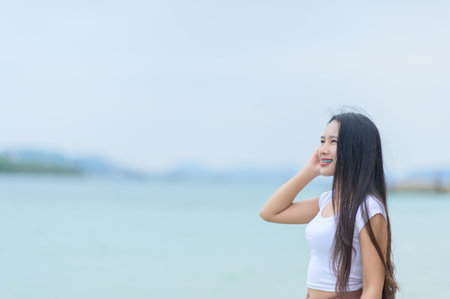 A woman in a white shirt and brown skirt enjoys a relaxing summer day at the beach, smiling and embracing the gentle breeze by the serene blue sea.の写真素材