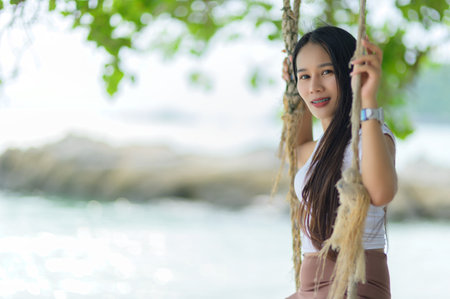 A woman wearing a white shirt and brown skirt enjoys a sunny summer day at the beach while swinging gently. The vibrant surroundings create a perfect backdrop for relaxation.の写真素材