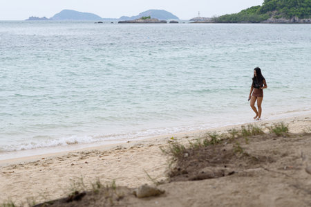 A solitary woman strolls along a serene beach, feeling the gentle waves lap against her feet. The picturesque landscape showcases lush greenery and distant islands.の写真素材