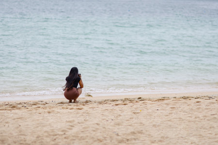 A serene scene featuring a woman squatting on the soft beach, focused on capturing the beauty of ocean waves. The tranquil waterfront invites relaxation and inspiration.の写真素材