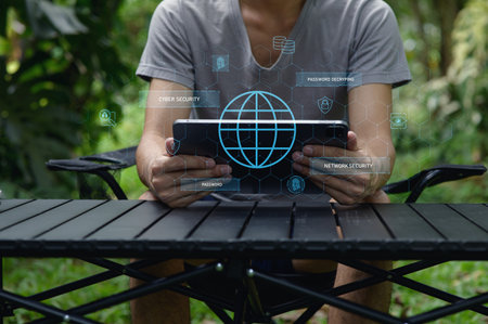 A man sitting outdoors holds a tablet showcasing network security graphics. This image highlights the importance of cybersecurity and digital safety in today's technology-driven world, nestled in a serene natural setting.の写真素材
