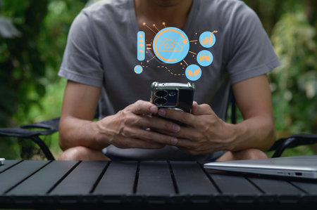 A young man sits outdoors, engaged with his smartphone while digital icons float above, symbolizing...