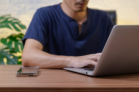 A person focused on a laptop, typing away with a smartphone beside them on a wooden desk, surrounded by a blurred plant, showcasing a modern work environment.の写真素材