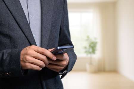 A businessman in a stylish suit holding a smartphone, standing in a modern office environment with soft lighting and a relaxed atmosphere. Perfect for representing modern communication.の写真素材
