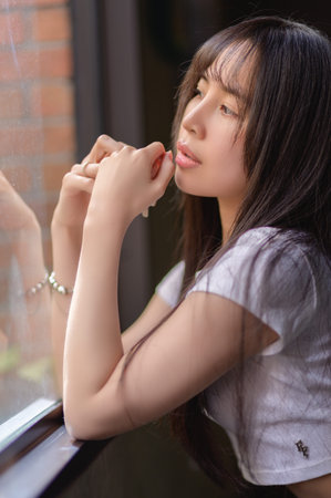 A serene portrait of a young woman gazing thoughtfully out of a window, bathed in natural light. The...
