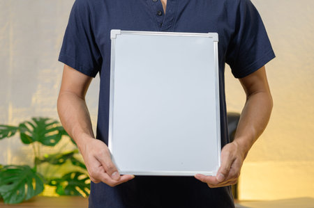 A person holds a blank whiteboard in a spacious indoor workspace. Lush green plants are visible in...