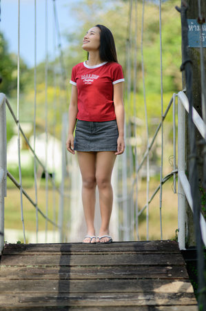 A young woman stands confidently on a wooden suspension bridge, embracing the beauty of nature. Her casual outfit reflects a joyful and carefree spirit in a sunny landscape.の写真素材
